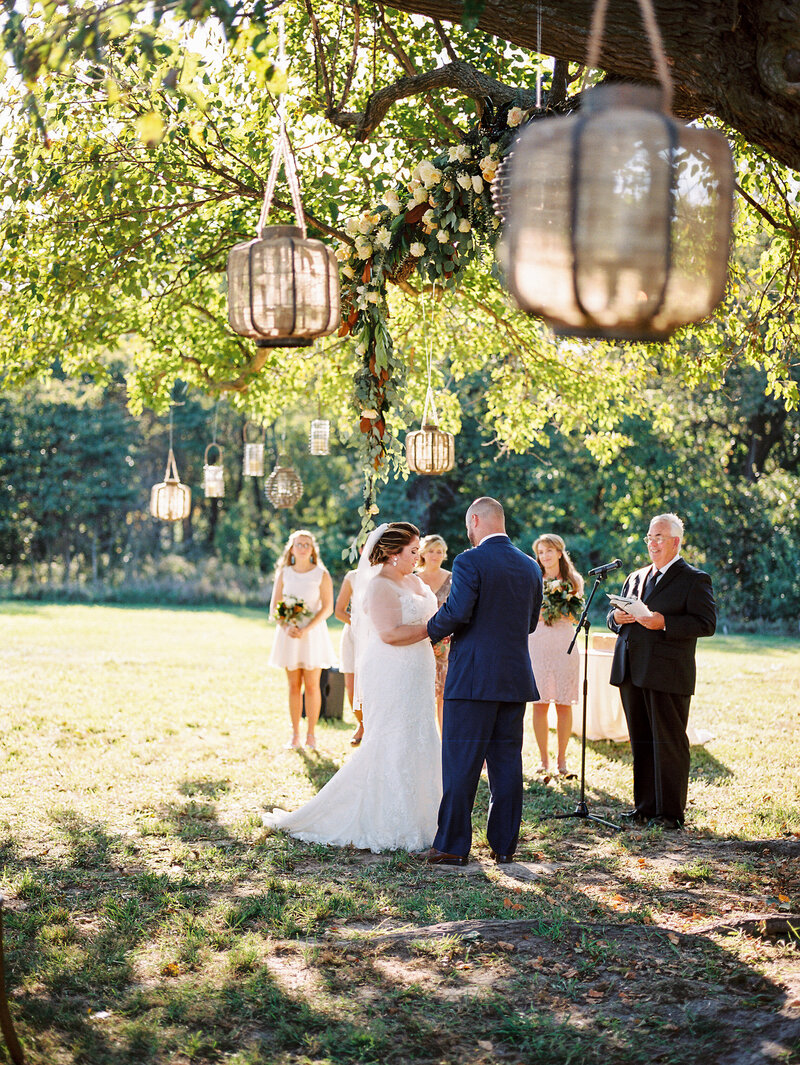 An intimate outdoor wedding ceremony under hanging lanterns with dappled sunlight filtering through the trees.