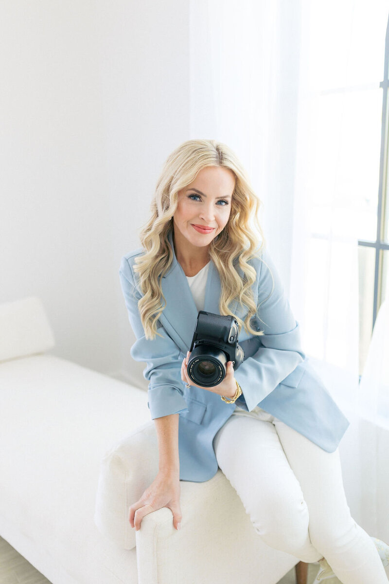 Woman with blonde hair and blue jacket holds film camera on white couch