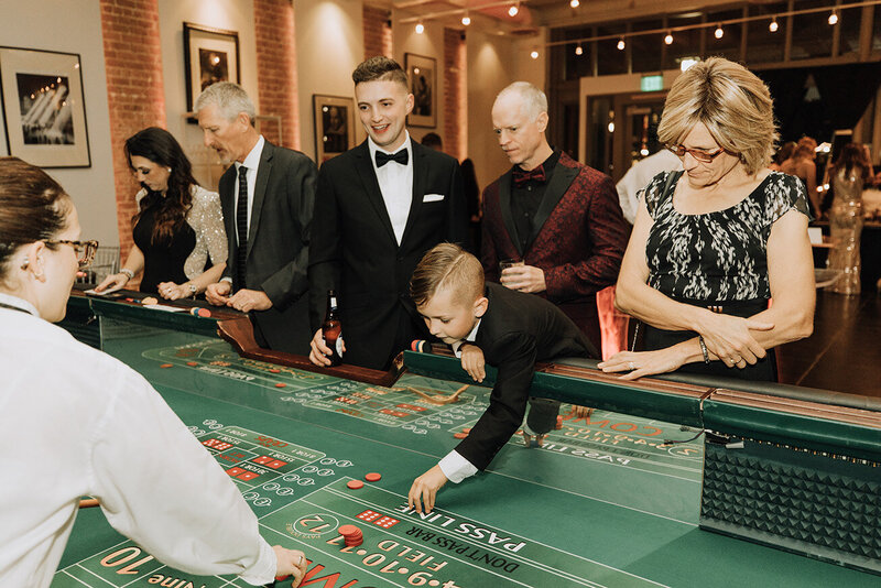 Guests dressed in formalwear enjoy a casino-themed reception at SoHo63, a Chandler wedding and event venue. A young boy enthusiastically reaches across the craps table as adults look on, showcasing the playful and customizable celebration options available at the venue.