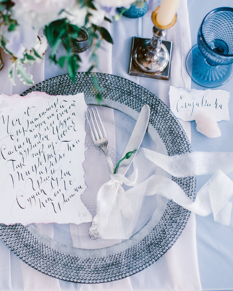 Beautiful wedding calligraphy cards sit atop a silver charger  plate with cutlery on luxury wedding table.
