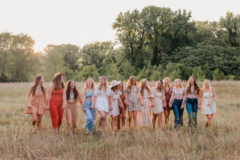 Rachel B Photography's Senior Rep Team of 14 seniors walk hand in hand in an open field  while smiling at one another.