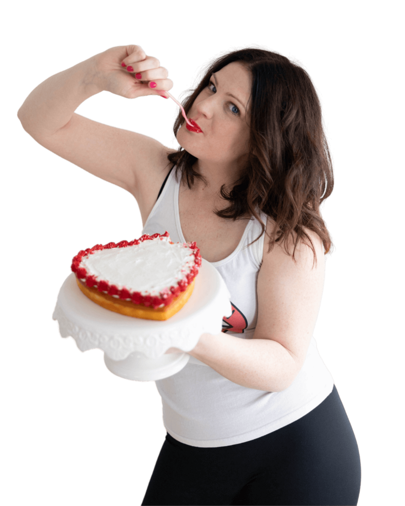 Emily wearing a white tank top and red lipstick, holding a fork in her mouth in one hand and a heart shaped cake in another.