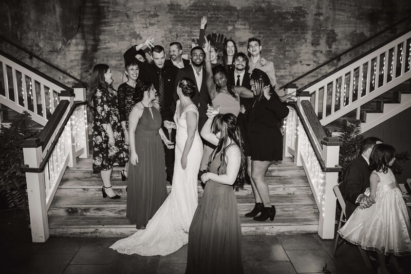 A black-and-white photo of a wedding party gathered on a staircase. The bride and groom stand in the center, surrounded by smiling guests posing playfully. Some people are laughing.