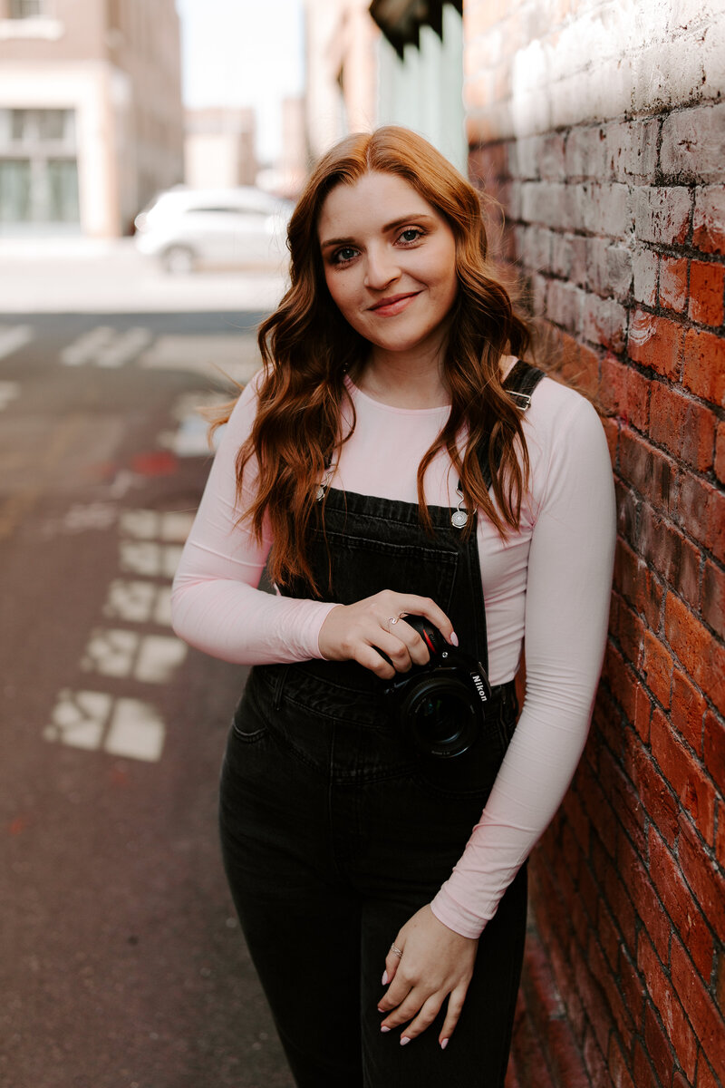 A portrait of the wedding and couples photographer while she smiles at the camera and leans against a brick wall in Downtown Spokane, Washington.