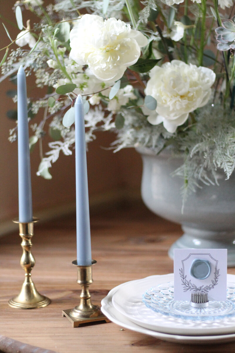A wedding floral arrangement of white peonies in a blue ceramic urn rests on a wooden table beside blue taper candles and ornate vintage plates with a wax seal placecard featuring a floral crest