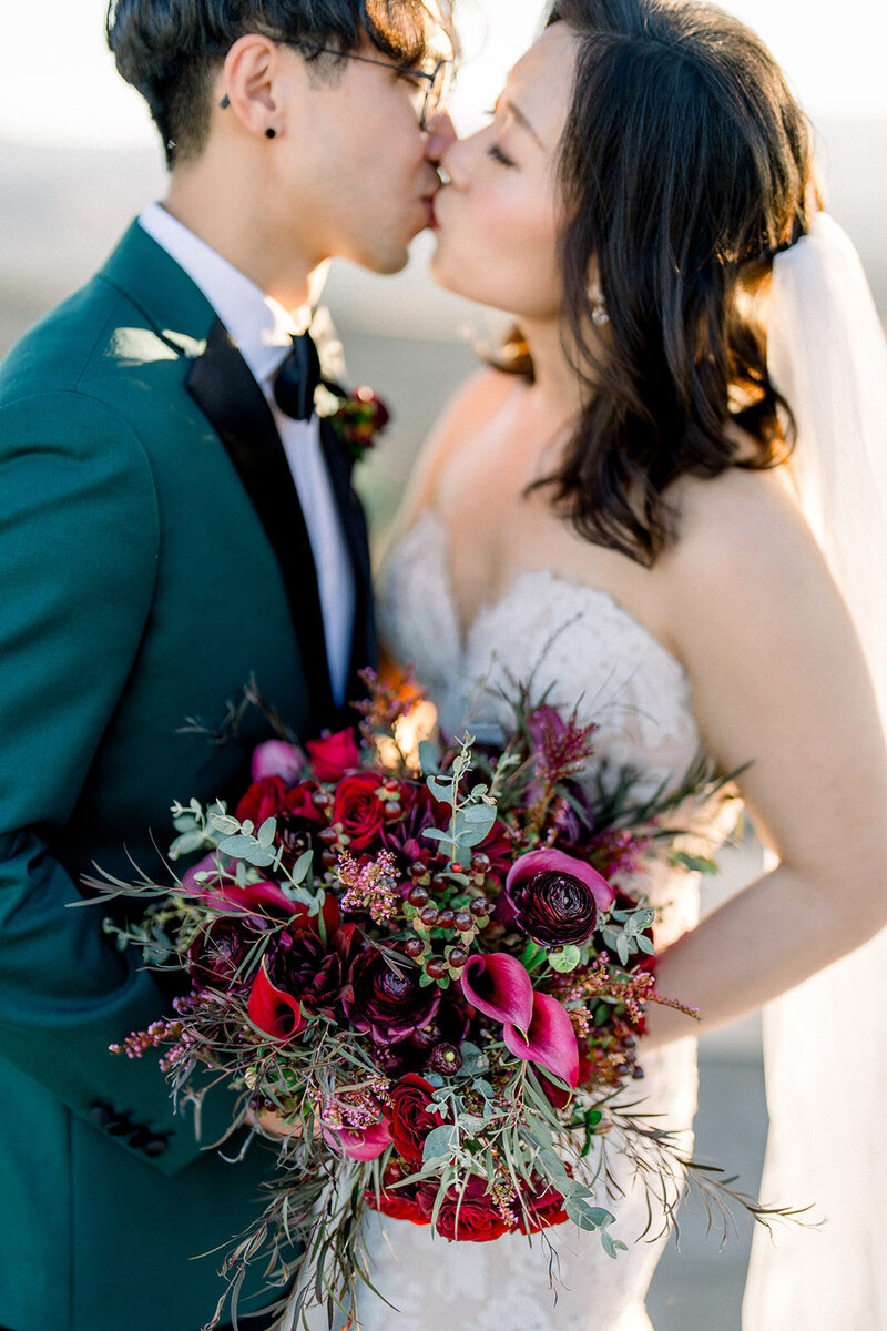 This stunning close-up captures a newlywed couple sharing a kiss, surrounded by the vivid colors of California’s golden light. The bride holds a striking bouquet of deep reds and purples, featuring roses, calla lilies, and eucalyptus. The groom’s dark teal suit contrasts beautifully with the floral arrangement, adding a modern yet romantic touch to this classic moment. The setting sun adds a soft, natural glow, enhancing the intimate atmosphere of this unforgettable California wedding.