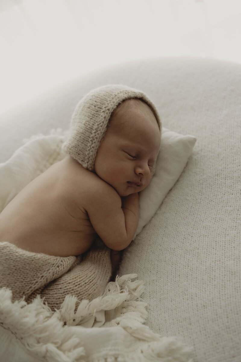 A sleeping baby lies on a cream-colored pillow, wearing a white knitted outfit, peacefully folded hand under cheek.