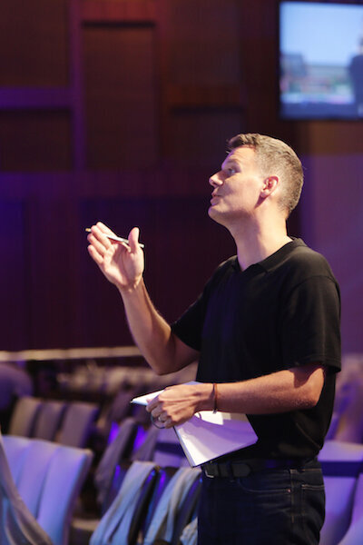 Man in a black shirt speaking and gesturing while holding papers in a dimly lit room.