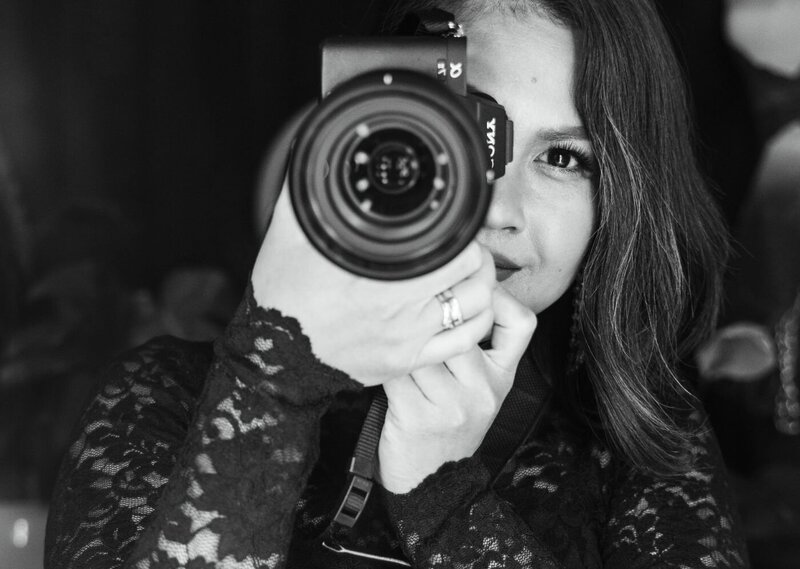 Black-and-white close-up of a woman photographer wearing a black lace top, looking through the viewfinder of her Sony camera while taking a picture.