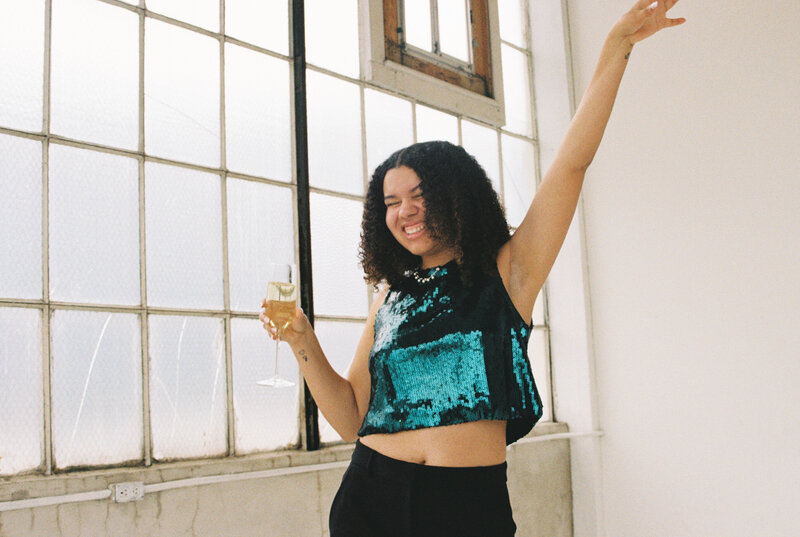 Photo of Catryce cheering in a teal top with a glass of mimosa with a window background