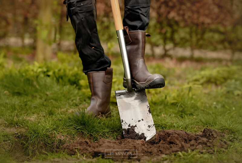Een man met een werkbroek en werkschoenen aan staat met een voet op de schop en de schop staat in het zand, met op de achtergrond gras