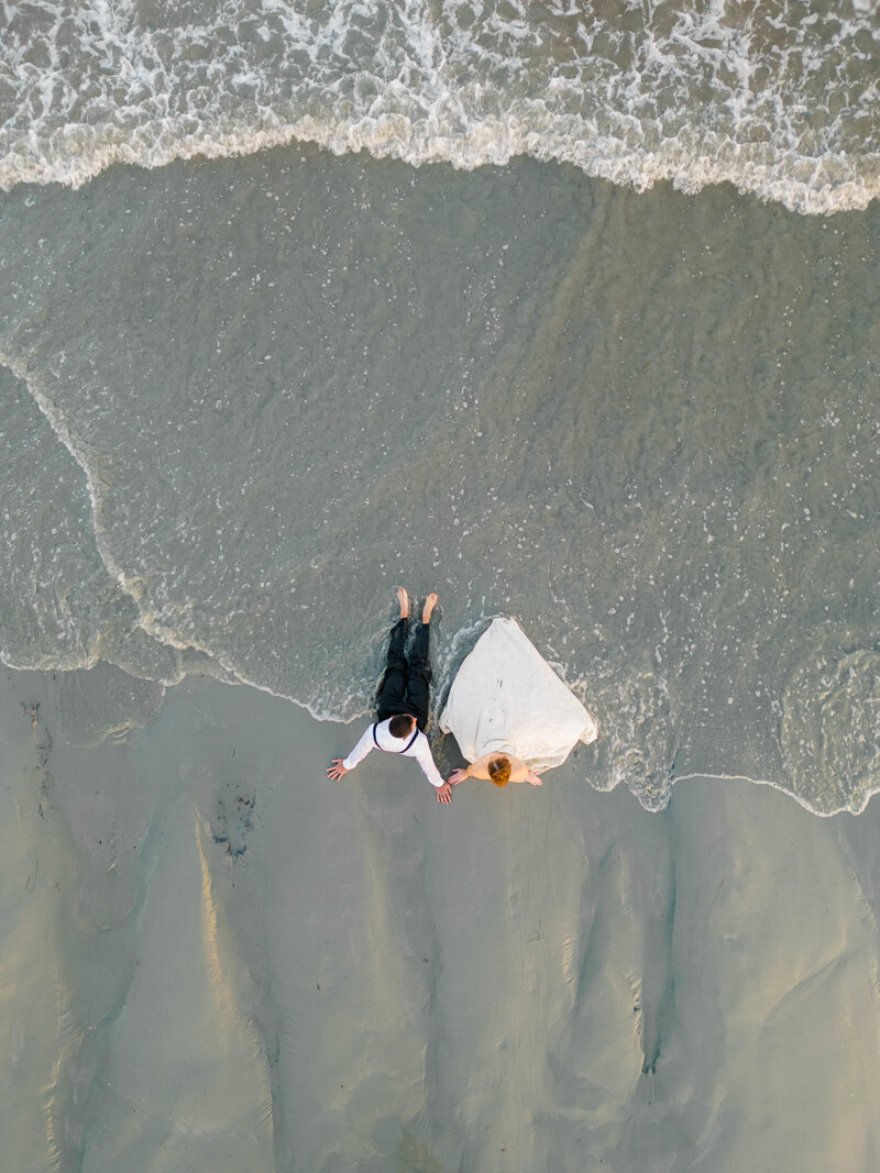 Rhode Island Elopement | Aerial view of a couple lying on wet sand with gentle waves touching them. The bride wears a white gown, and the groom is in a black suit.