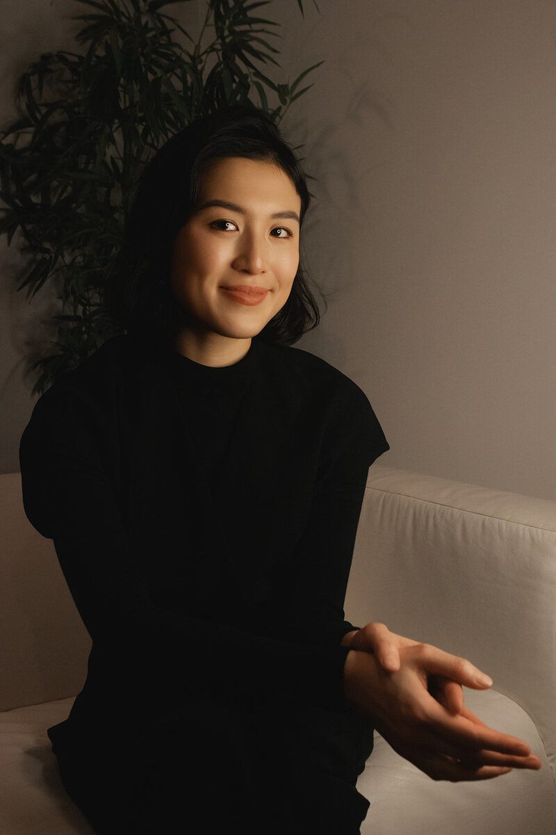 Woman with short dark hair weating a black long-sleeved shirt  softly smiles at camera with her palms together while sitting on an off-white couch with a tree behind her.