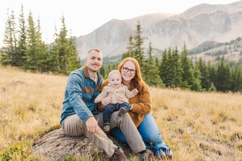 Family of 3 joyfully posing together on a rock at their family session in the Snowy Range Mountains near Laramie, Wyoming. 