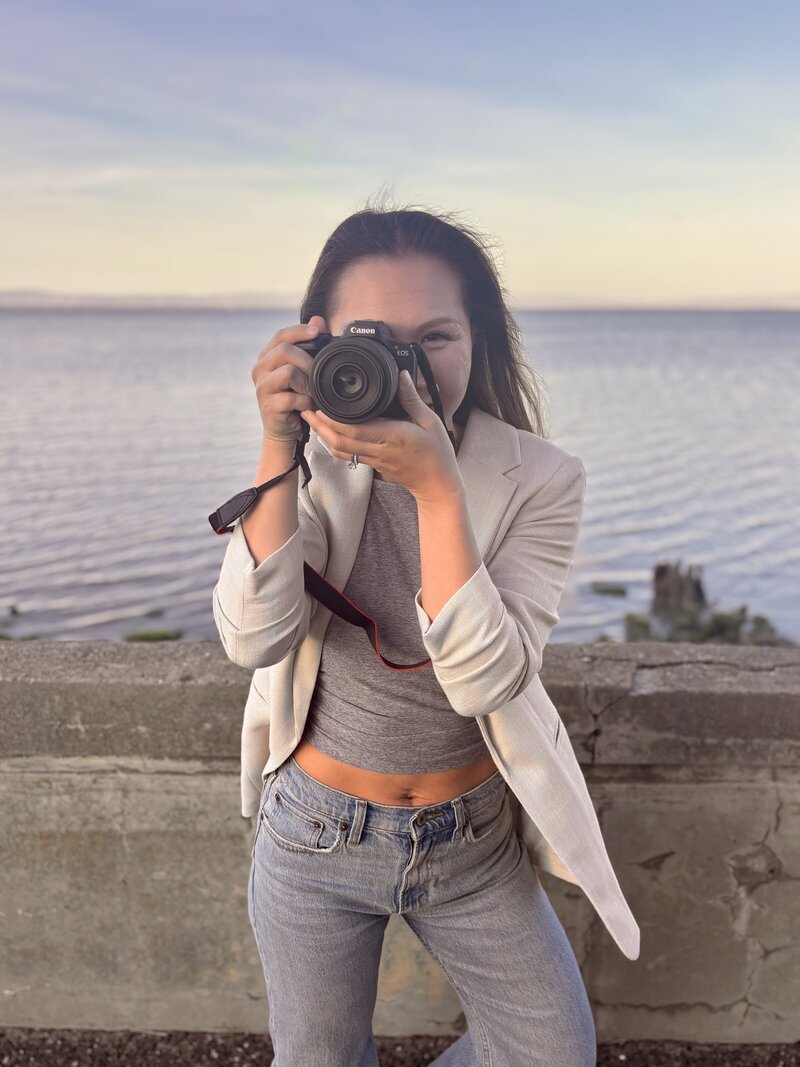 Ellen, owner of Ellobelle Photography, relaxed and smiling near ocean backdrop holding canon mirrorless camera– San Francisco Peninsula family and newborn photographer.