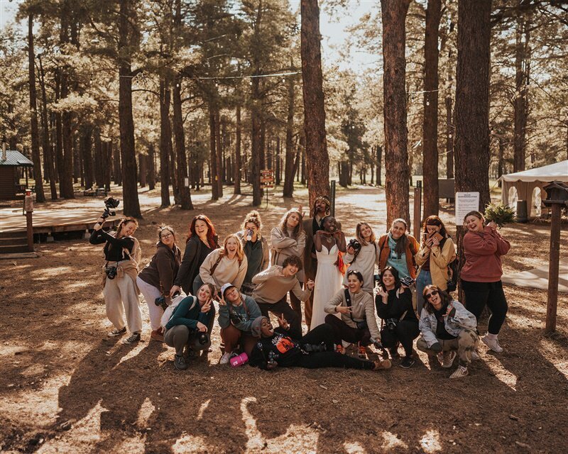 A group of photographers, at Aglow, gather in a wooded area, posing playfully together. The scene is filled with camaraderie and laughter among the tall trees and warm sunlight.