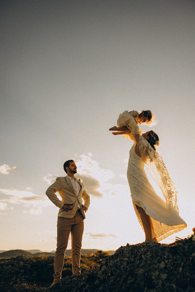 Natural elopement photography capturing a bride holding her toddler in the air while the groom watches, during sunset on Emu Mountain Sunshine Coast