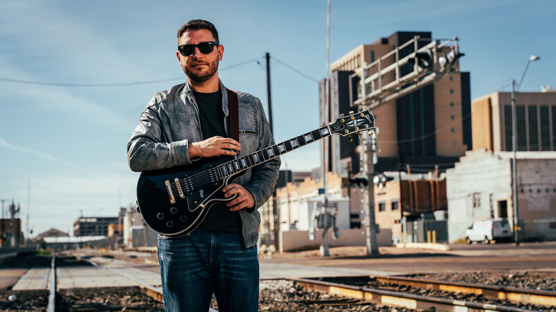 A man wearing sunglasses, a gray leather jacket, and jeans stands confidently with a black electric guitar beside railroad tracks. Behind him, an industrial setting with buildings and equipment provides a gritty, urban backdrop. His casual pose and direct gaze lend a cool, relaxed vibe to the image.