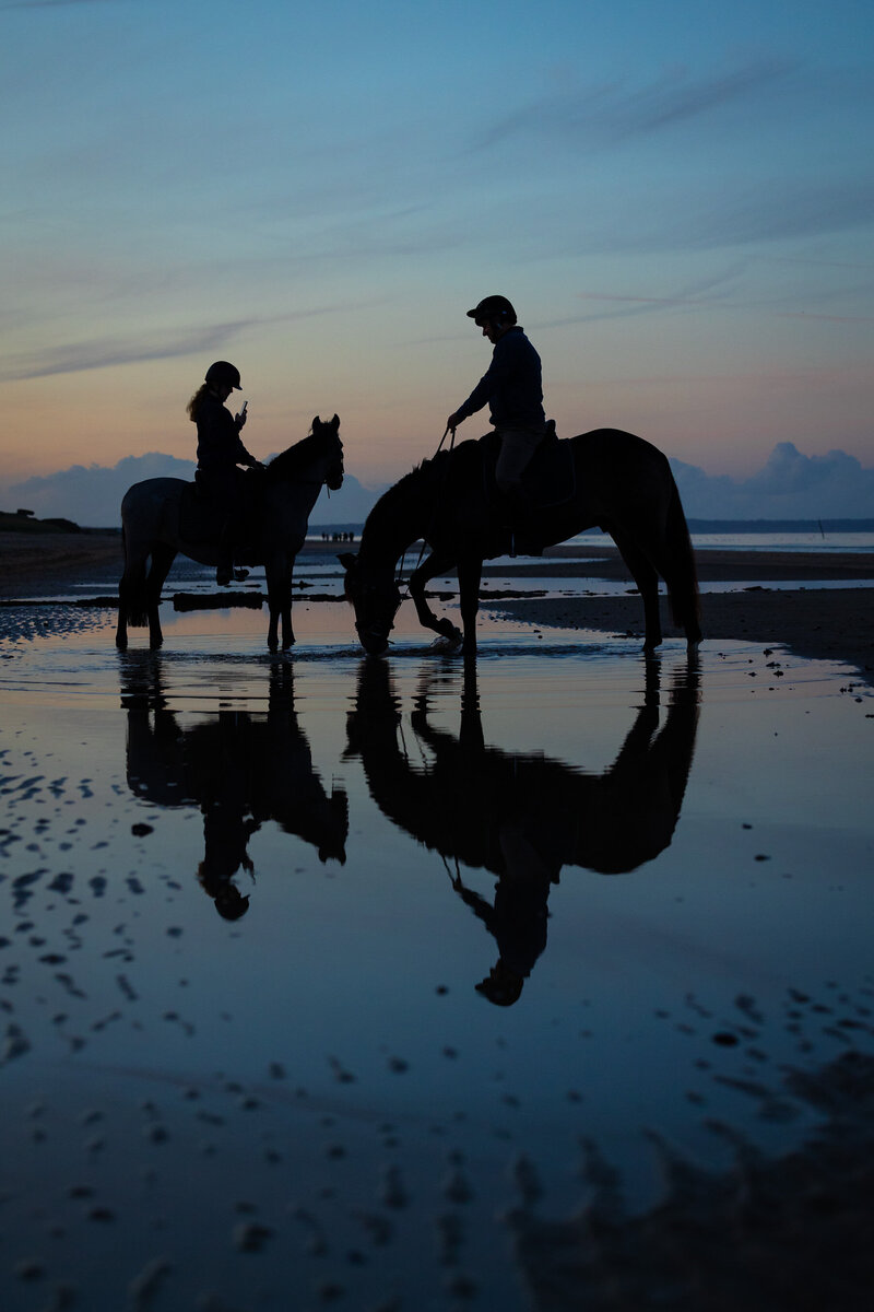 A silhouette of a father and daughter sitting on s horse and pony on a beach at sunset with their reflection showing in a pool of water