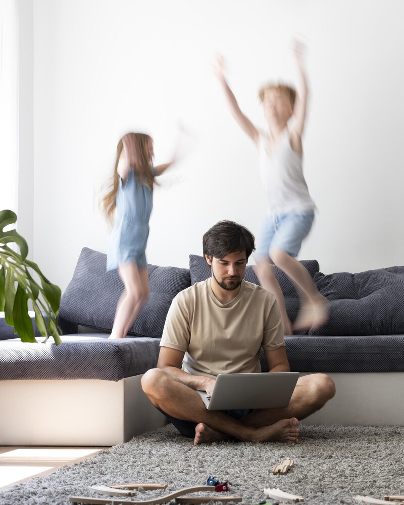 Father trying to work from home while children play in the background, illustrating the overwhelm of modern parenting and the need for somatic support in New York City.
