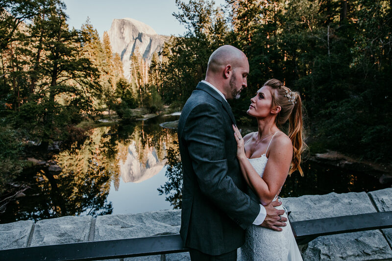 Bride and groom sharing an intimate moment on a bridge in Yosemite National Park, with Half Dome reflecting in the river at sunset.