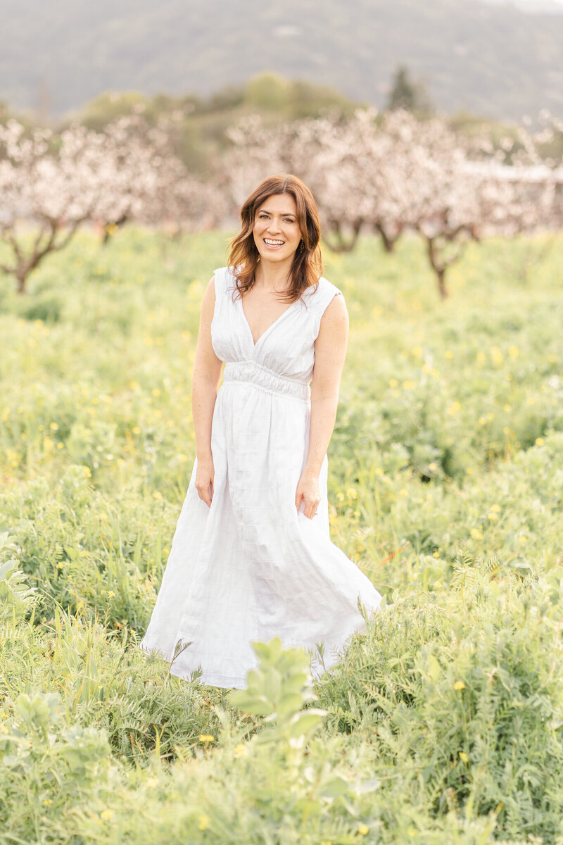 woman standing in a bay area orchard in a field of mustard wearing a white dress and smiling at the camera