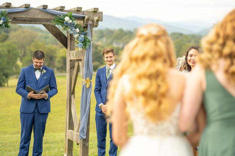 Bride walking down the aisle towards her groom at Renback Barn, with the groom smiling under a floral-adorned wooden arch, set against the scenic Virginia mountains.