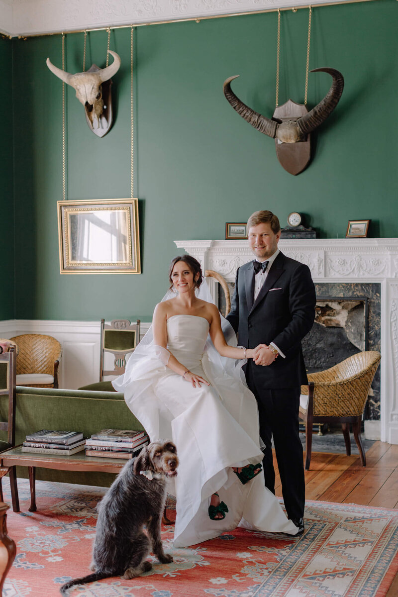 A bride in a strapless gown and groom in a black tuxedo stand with a dog  at Islay House, during their Scotland destination wedding planned by ByChenai..