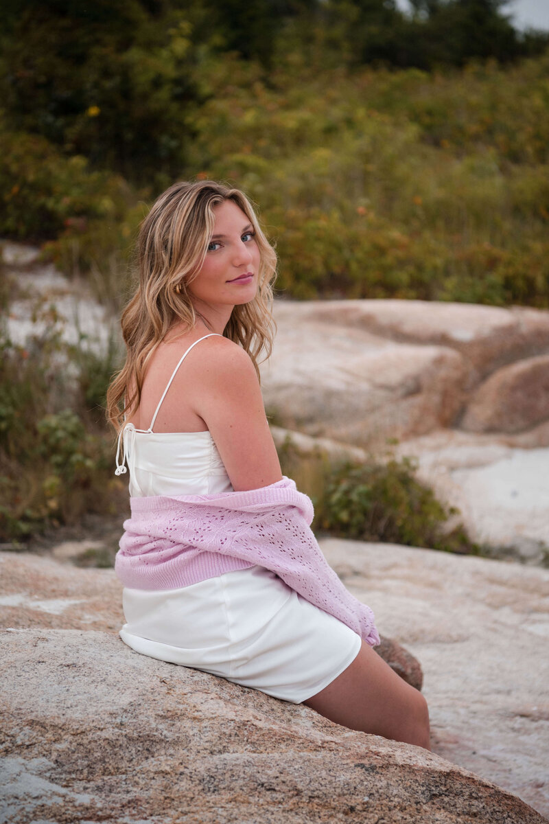 A senior sits on a rock and smiles slightly at the camera with Wingaersheek beach grass in the background.