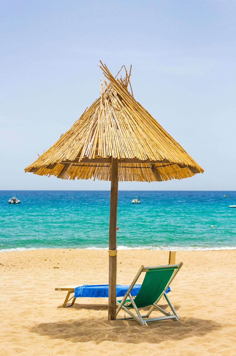 A serene beach scene featuring a rustic thatched umbrella shading a green reclining chair and a blue towel, set on golden sand with the turquoise ocean in the background. This idyllic setting embodies the relaxation and escape offered by vacation fundraising for charities.