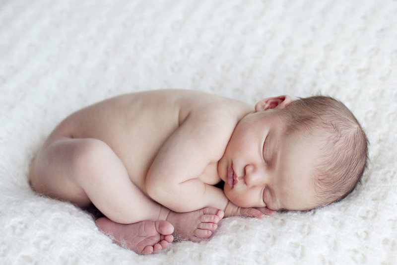 A newborn baby sleeps curled up on a white blanket