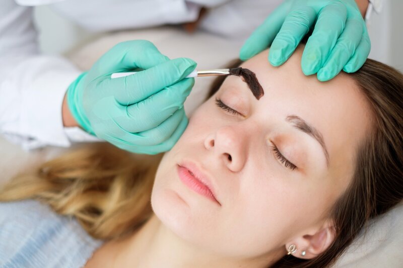 Closeup image of a woman lying down getting her eyebrows tinted from an esthetician wearing aqua blue gloves