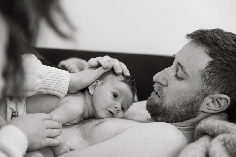 A black and white image capturing a joyful moment between a father and his young son. The dad is playfully throwing a blanket over his head while the baby laughs with delight