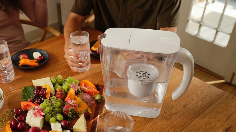 Clearly Filtered water pitcher sitting on a table with glass of water and fruit and cheese platter
