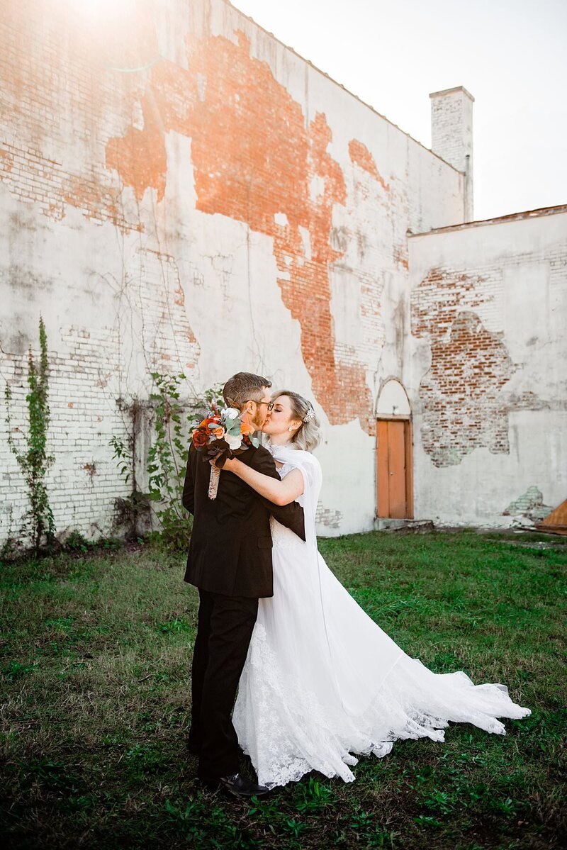 Couple kissing on their wedding day, she's in her moms vintage dress and there is a white and exposed brick wall behind them