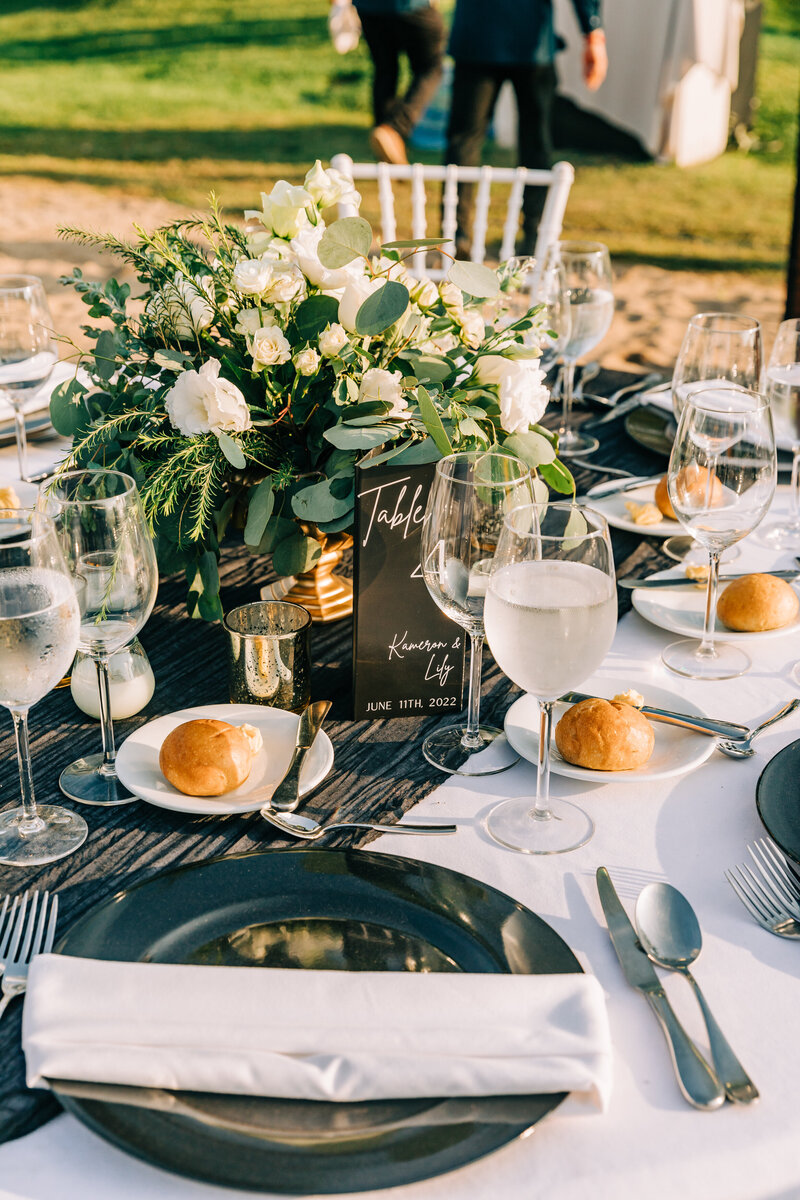 A beautifully arranged outdoor wedding reception table featuring a floral centerpiece of white roses and eucalyptus. The table is set with glasses, bread rolls, and a black table card that reads ‘Table 4.