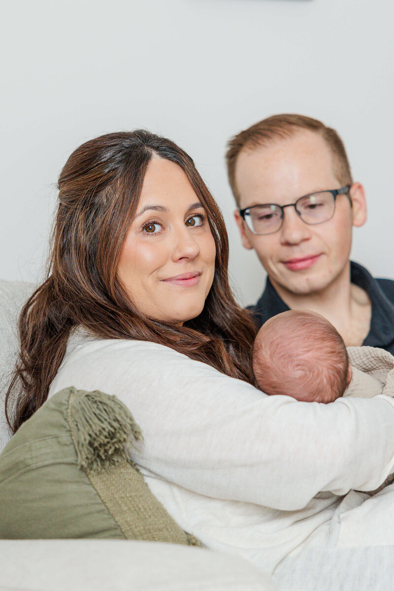 Mom and dad with their newborn sitting on a beige couch in the living room. Dad is looking at the baby while mom smiles at the camera during a lifestyle in-home newborn session in Southeast Michigan.