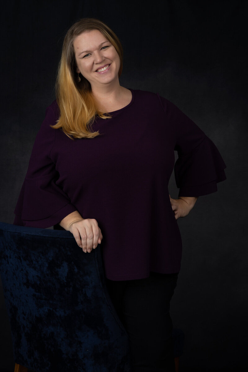 photographer-headshot-wearing-purple-shirt-in-studio-with-hand-on-hip-holding-blue-chair-in-arlington-tx