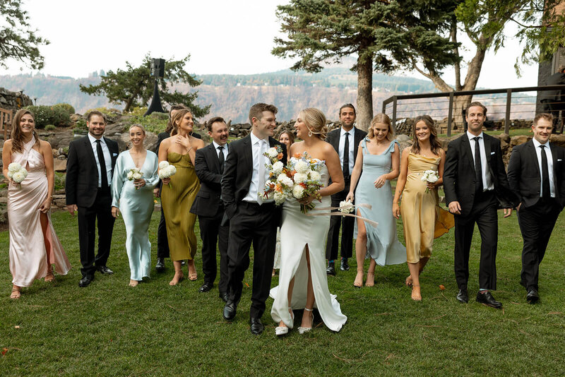 A group wedding photograph. The bride and groom are in the middle of the  photo. They are looking at each other smiling. The bride is wearing a strapless white wedding dress and holding a large wedding bouquet. The groom is wearing a black suit with a white neck tie. Behind the couple, they are surrounded by their wedding party who are all walking and smiling. The men are wearing black suits with black ties, and the women are each wearing a different color bridesmaid dress. Everyone is walking along green grass, and in the background you can see a cliff, mountains, and trees. The setting is at The Griffin House wedding venue in Hood River, Oregon.