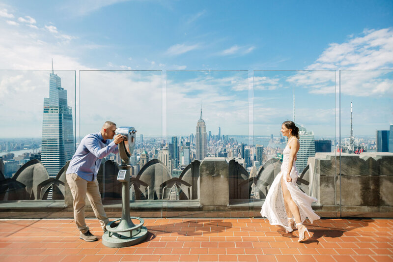 A person looking through a viewfinder towards their partner while they are on the top of a skyscraper.