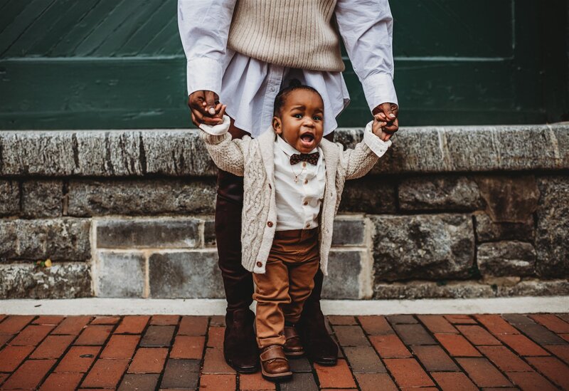 fall family pictures with father holding his toddlers hands as he helps him to walk captured by Family photographers Maryland