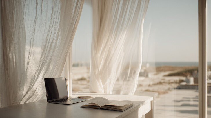 A sunlit workspace overlooking the beach with sheer curtains, an open book, and a laptop, symbolizing creativity and flow within Kathalyst Design Studio’s design process.