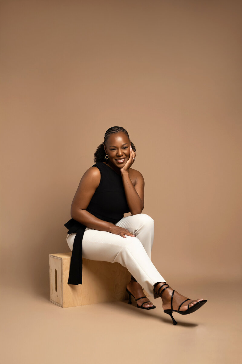 Professional headshot of Rianne Nycole, a smiling Black woman with braided hair in jeans and a white button-down shirt, sitting against a light background.
