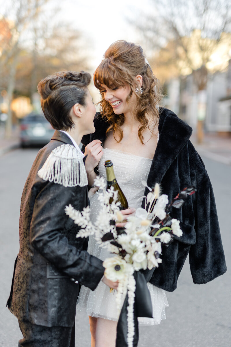 Nonbinary newlyweds smiling and staring at one another in the street in Georgetown DC while holding a bouquet and champagne bottle