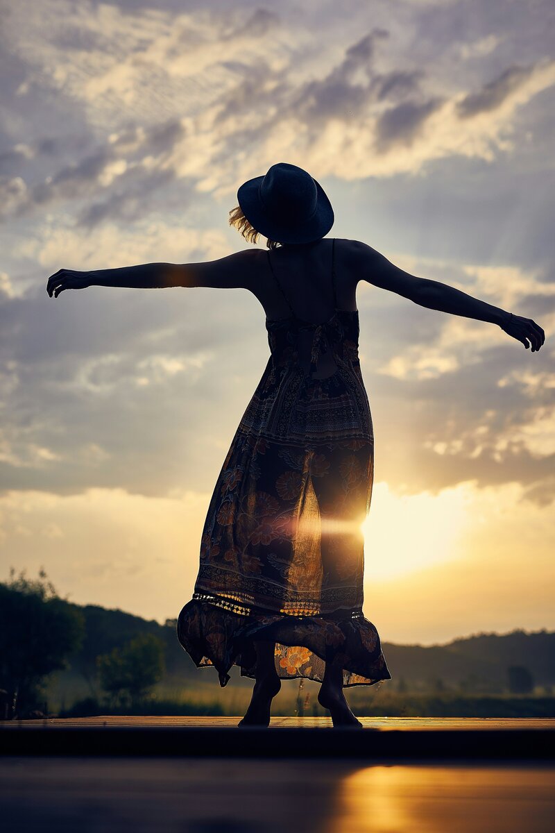 Image of a femme presenting person with their arms stretched out during sunset while wearing a dress and top hat