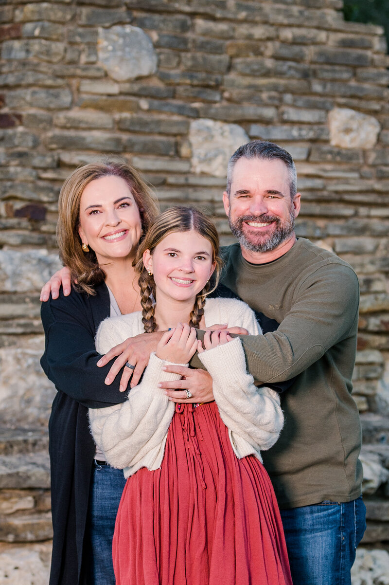 Family of 3 having a portrait taken with a stone wall. Photo taken by Dripping Springs Texas based photographer Lydia Teague.