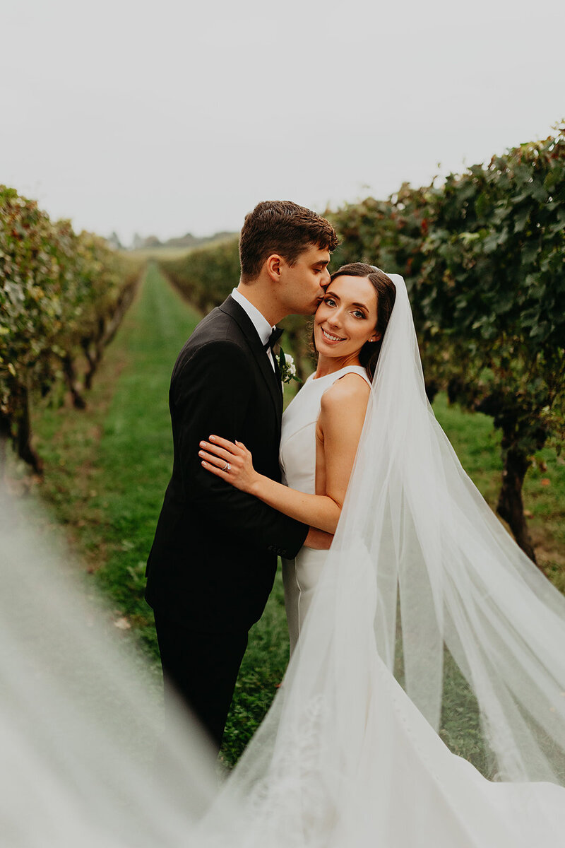 The groom plants a tender kiss on the bride's cheek, her veil gracefully unfolding towards the camera, capturing the romantic moment at the vineyard.