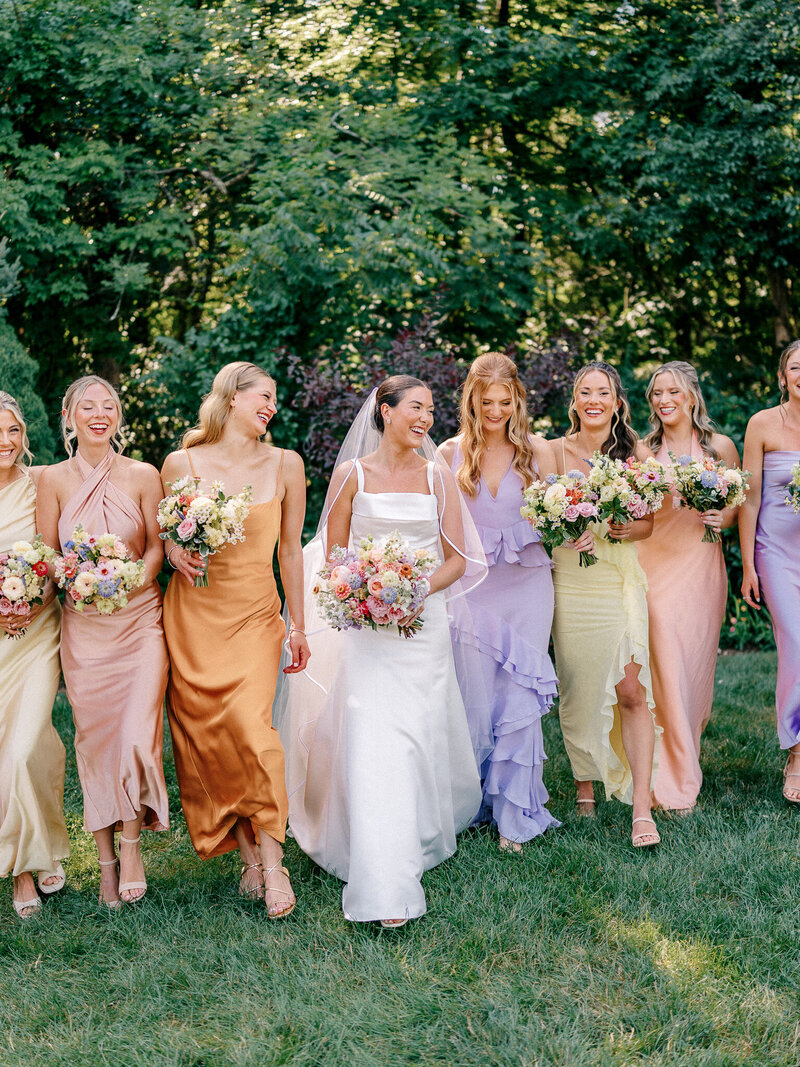 Bride and bridesmaids in pastel dresses walking in a line and laughing outside during bride's New England wedding.