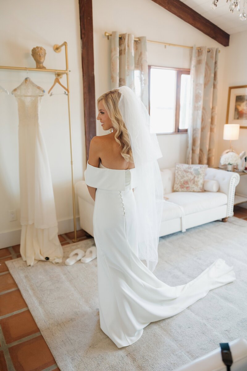 A bride in a white dress holds a bouquet while standing with and kissing the groom in a tuxedo under a stone archway surrounded by greenery.