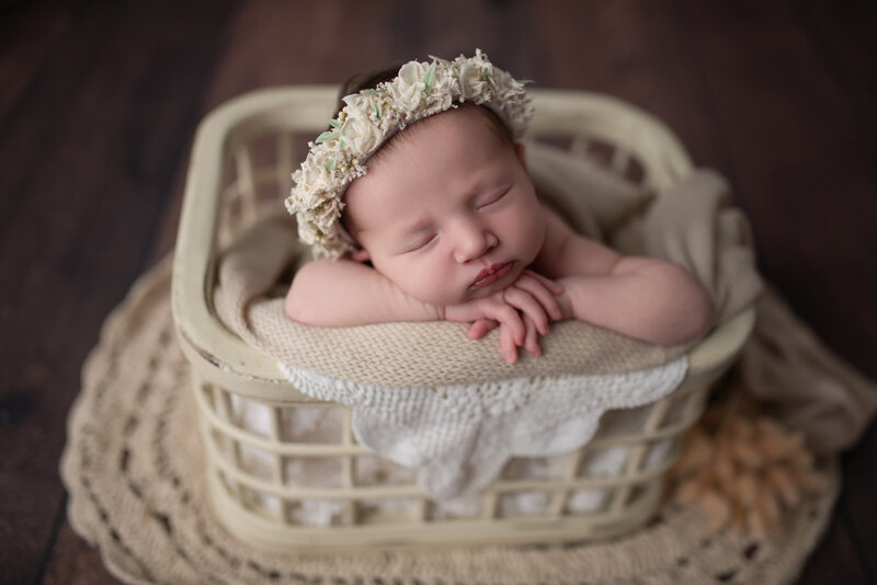 Styled in neutrals and vintage lace, this floral-crowned newborn girl rests gently in a cream basket pose.